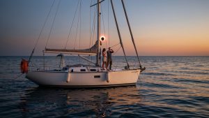 Voilier blanc au mouillage sur une mer calme au crépuscule, avec deux femmes et un homme observant une éclipse solaire à travers des lunettes de protection, devant un horizon ouest dégagé.